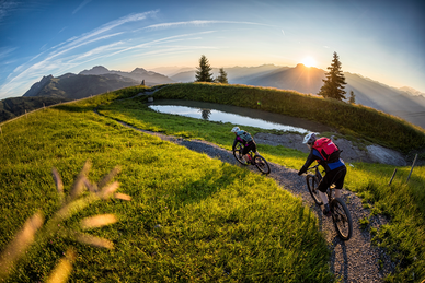 Zwei Mountainbiker fahren bei Sonnenuntergang auf einem schmalen Weg durch grüne Wiesen mit Blick auf das Großarltal. | © mtb_travel