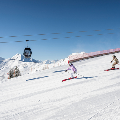 Two skiers descend a groomed slope in Grossarltal beneath a gondola lift with snowy mountains and blue sky | © Lorenz Masser