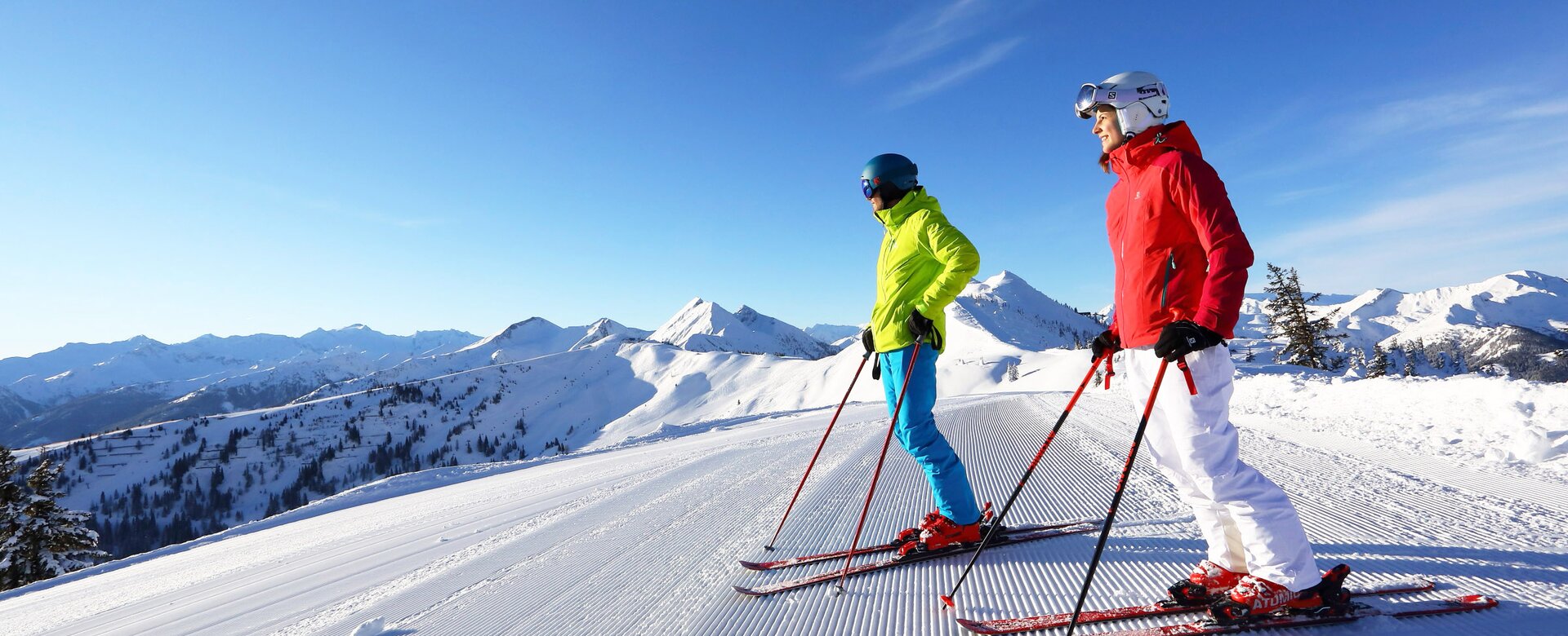 Two skiers standing on a freshly groomed slope looking down, snow-capped mountains and a bright blue sky with sun in the background | © Tourismusverband Großarltal
