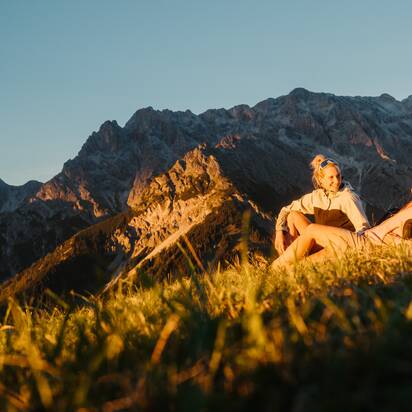Two women sit on a grassy slope in front of a rocky mountain massif in warm evening light in the Hochkönig region. | © Hochkönig Tourismus GmbH