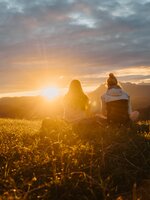 Zwei Frauen sitzen auf einer Wiese und blicken in den Sonnenuntergang über Bergsilhouetten unter wolkigem Himmel. | © Hochkönig Tourismus GmbH