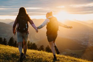 Zwei Frauen mit Rucksäcken gehen über eine Wiese im Abendlicht mit Blick auf Berge und Tal in der Region Hochkönig. | © Hochkönig Tourismus