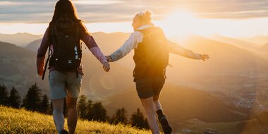 Two women with backpacks walk across a meadow in evening light overlooking mountains and valley in the Hochkönig region. | © Hochkönig Tourismus