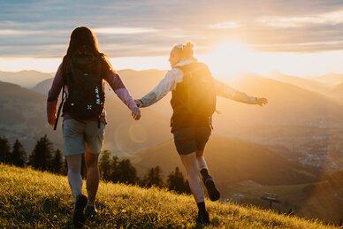 Two women with backpacks walk across a meadow in evening light overlooking mountains and valley in the Hochkönig region. | © Hochkönig Tourismus