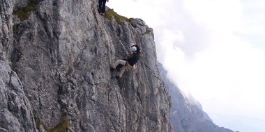 Zwei Kletterer an steiler Felswand gesichert, einer steigt, der andere sichert, Wolken und Bergpanorama im Hintergrund
