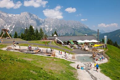 Children play at water features and playground at Toni’s Alm, with a mountain hut, terrace, and alpine peaks in the background | © Gruber Michael