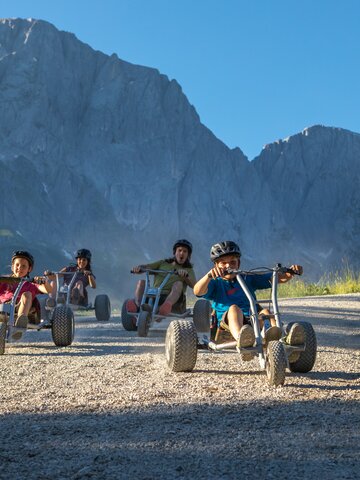 Kids race downhill on mountain carts with dramatic Hochkönig peaks behind, wearing helmets, sunny day | © Roland Haschka
