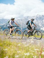 Two mountain bikers ride on a gravel path with steep rocky mountains and green alpine meadows at Hochkönig. | © Hochkönig Tourismus GmbH