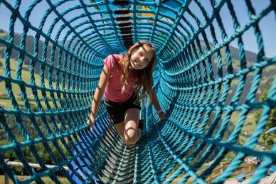 Mädchen krabbelt lachend durch blauen Klettertunnel am Schlossspielplatz Natrun, Almwiese im Hintergrund | © Roland Haschka