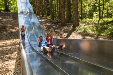 Familie rutscht lachend auf breiter Metallrutsche im Waldrutschenpark durch den schattigen Wald bei Maria Alm | © Roland Haschka