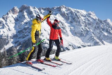 Zwei Skifahrer in bunter Kleidung geben sich High-Five auf Skipiste vor Bergkulisse | © Hochkönig Tourismus GmbH