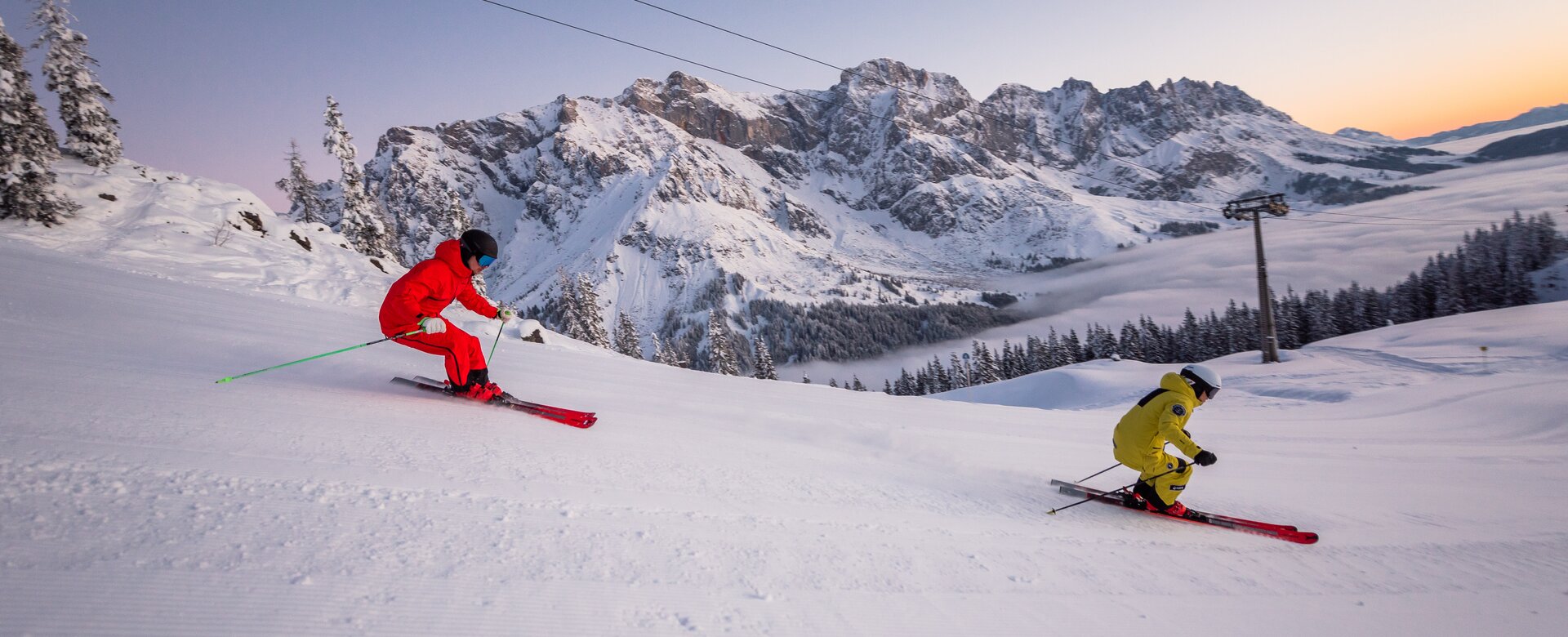 Ein Skifahrer in gelbem Skianzug und einer in rotem fahren die Piste hinunter und im Hintergrund geht die Sonne gerade auf | © Hochkönig Tourismus