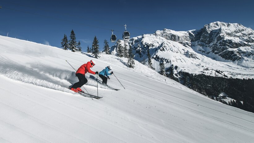 Zwei Skifahrer auf der Piste mit Gondel im Hintergrund | © Hochkönig Tourismus GmbH