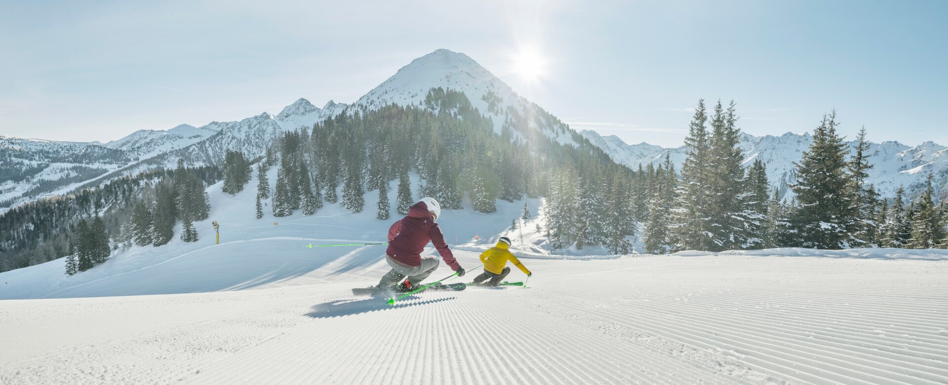 Zwei Skifahrer fahren die frisch präparierte Piste hinunter und es sind schneebedeckte Berge zu sehen | © Peter Burgstaller