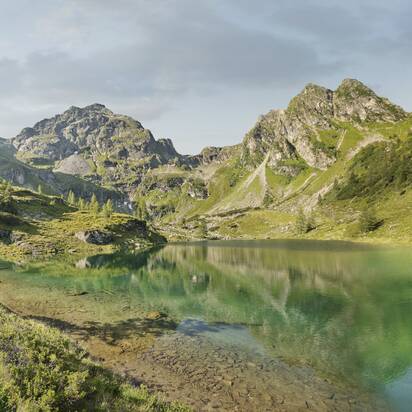 Two hikers walk along a turquoise mountain lake, surrounded by alpine meadows, trees and rugged mountain peaks | © Peter Burgstaller