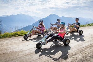 Four helmeted riders ride mountain go-karts along a wide gravel road with mountains in the background | © Planai, Harald Steiner