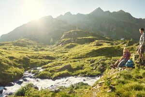 Two hikers rest by a mountain stream in the sun with green alpine pastures and mountain peaks in the background | © Peter Burgstaller