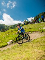 A child wearing a helmet rides a mountain bike on a winding trail across a green alpine meadow, people behind | © Christine Höflehner