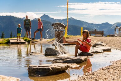 Ein Mädchen sitzt auf Steinen im flachen Wasser und spritzt, während eine Familie vor Bergkulisse steht | © Planai, Christine Höflehner