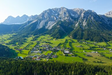Luftaufnahme des Ramsauer Plateaus mit weiten grünen Wiesen, kleinen Häusern und dem imposanten Dachsteinmassiv im Hintergrund | © Herbert Raffalt