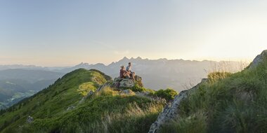Two hikers sit on a rock and enjoy the mountain view during sunrise on the Reiteralm | © Peter Burgstaller