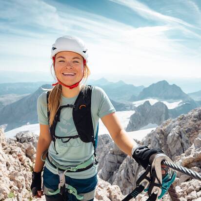 Kletterin mit Helm, Gurt und Handschuhen am Stahlseil zwischen hellem Fels und schneebedeckten Bergen am Dachstein | © Mathäus Gartner