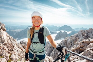 Kletterin mit Helm, Gurt und Handschuhen am Stahlseil zwischen hellem Fels und schneebedeckten Bergen am Dachstein | © Mathäus Gartner