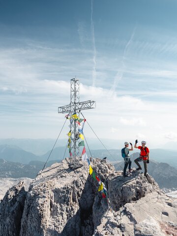 Two climbers wearing helmets stand beside a summit cross on a rocky peak above a wide alpine landscape | © Mathäus Gartner