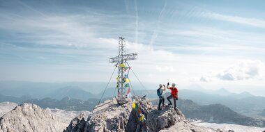 Two climbers wearing helmets stand beside a summit cross on a rocky peak above a wide alpine landscape | © Mathäus Gartner