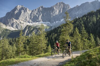Two mountain bikers ride along a gravel path through alpine terrain with forest and a bright rock face below a blue sky | © Lorenz Masser