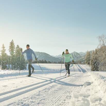 Zwei Langläufer in Sportkleidung gleiten bei Sonne auf gespurter Loipe durch verschneite Winterlandschaft | © Peter Burgstaller