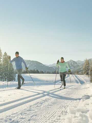 Two cross-country skiers in sportswear glide on sunny groomed trail through snowy winter landscape | © Peter Burgstaller