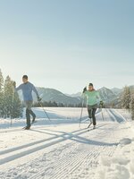 Two cross-country skiers in sportswear glide on sunny groomed trail through snowy winter landscape | © Peter Burgstaller