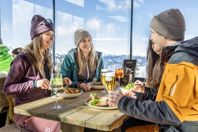 Four people in ski gear dining at a wooden table with mountain view in the restaurant | © Mirja Geh
