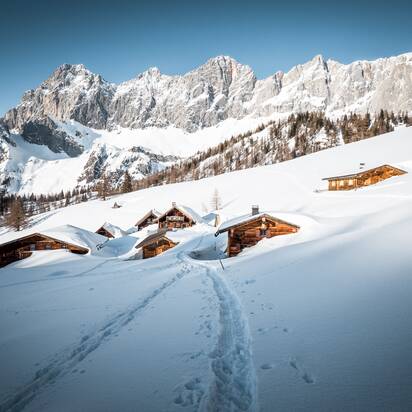Footprints lead to snow-covered alpine huts below Dachstein mountains in wintry alpine landscape | © Mathäus Gartner