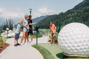 Eine Familie spielt Minigolf neben einer grossen Golfball-Skulptur vor Bergen und bewaldeten Hängen. | © JOSalzburg