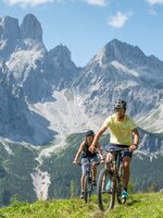 Zwei Radfahrer fahren über eine grüne Wiese in Filzmoos vor steilen grauen Felsbergen unter blauem Himmel | © Lorenz Masser