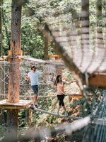 Zwei Personen balancieren auf einem Seil im Hochseilpark zwischen Plattformen und Netzen im Wald. | © JOSalzburg