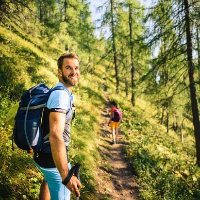 Man with backpack hikes through sunlit alpine forest trail, smiling back at camera; woman walking ahead in the background | © Salzburger Sportwelt - Christian Schartner