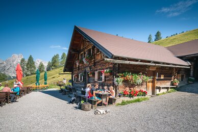 A family sits at a wooden table in front of a flower-adorned alpine hut in Filzmoos, surrounded by mountains and green meadows. | © Salzburger Sportwelt/Christian Schartner
