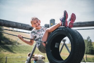 Lachendes Mädchen sitzt auf einer Reifenschaukel auf dem Spielplatz, hält sich an Ketten fest, Sommerwiese im Hintergrund | © Salzburger Sportwelt