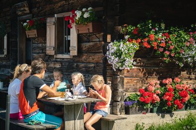 A family enjoys a snack at a wooden table in front of a rustic alpine hut decorated with colorful flowers in Filzmoos. | © Salzburger Sportwelt/Christian Schartner