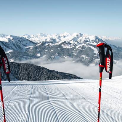 Two ski poles and red skis on freshly groomed ski slope with view of snow-covered alpine landscape | © Snow Space Salzburg Christian Schartner