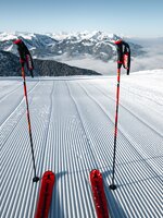 Zwei Skistöcke und rote Ski stehen auf frisch präparierter Skipiste mit Blick auf verschneite Alpenlandschaft | © Snow Space Salzburg Christian Schartner