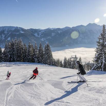 A family is skiing down the piste and the valley is covered in fog, while a bright blue sky can be seen above | © Michael Grössinger