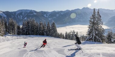 A family is skiing down the piste and the valley is covered in fog, while a bright blue sky can be seen above | © Michael Grössinger
