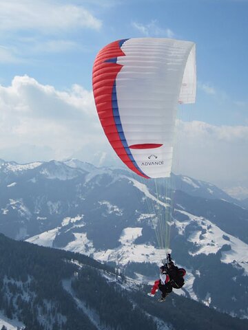 Paraglider with red, blue and white glider flies over mountains covered in forest and snowy pistes