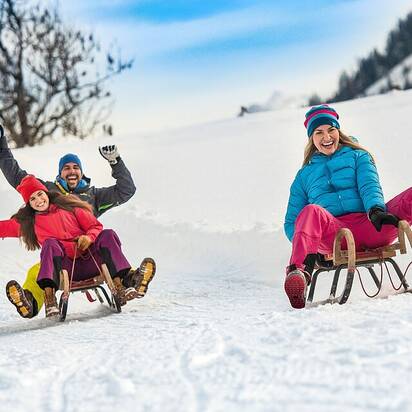 Drei Personen rodeln lachend auf einer präparierten Schneebahn, umgeben von winterlicher Landschaft. | © Flachau Tourismus