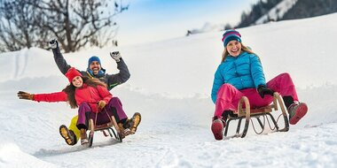 Drei Personen rodeln lachend auf einer präparierten Schneebahn, umgeben von winterlicher Landschaft. | © Flachau Tourismus