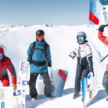 Four winter sports enthusiasts pose with giant ski passes in the snow before alpine mountain scenery.